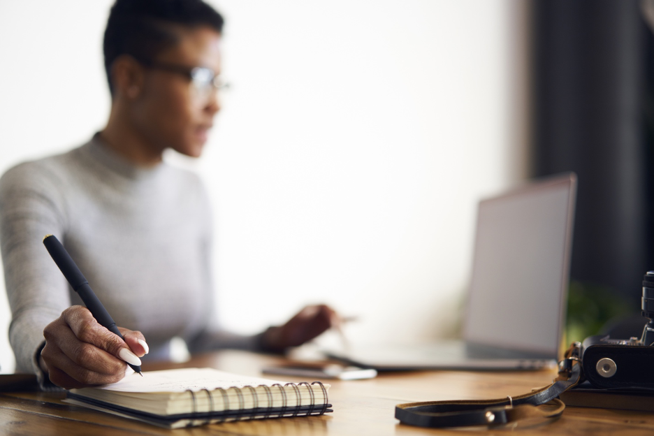 African American woman writing on notepad looking at computer